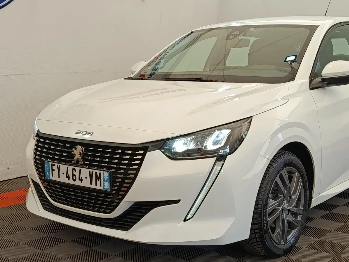Vue 3/4 avant droit d'une Peugeot 208 blanche de 2021 avec phares LED allumés et calandre noire distinctive.