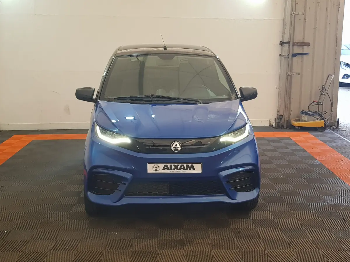 Vue frontale d'une AIXAM AMBITION 0.5 CITY SPORT bleu avec phares allumés et rétroviseur caméra côté conducteur.