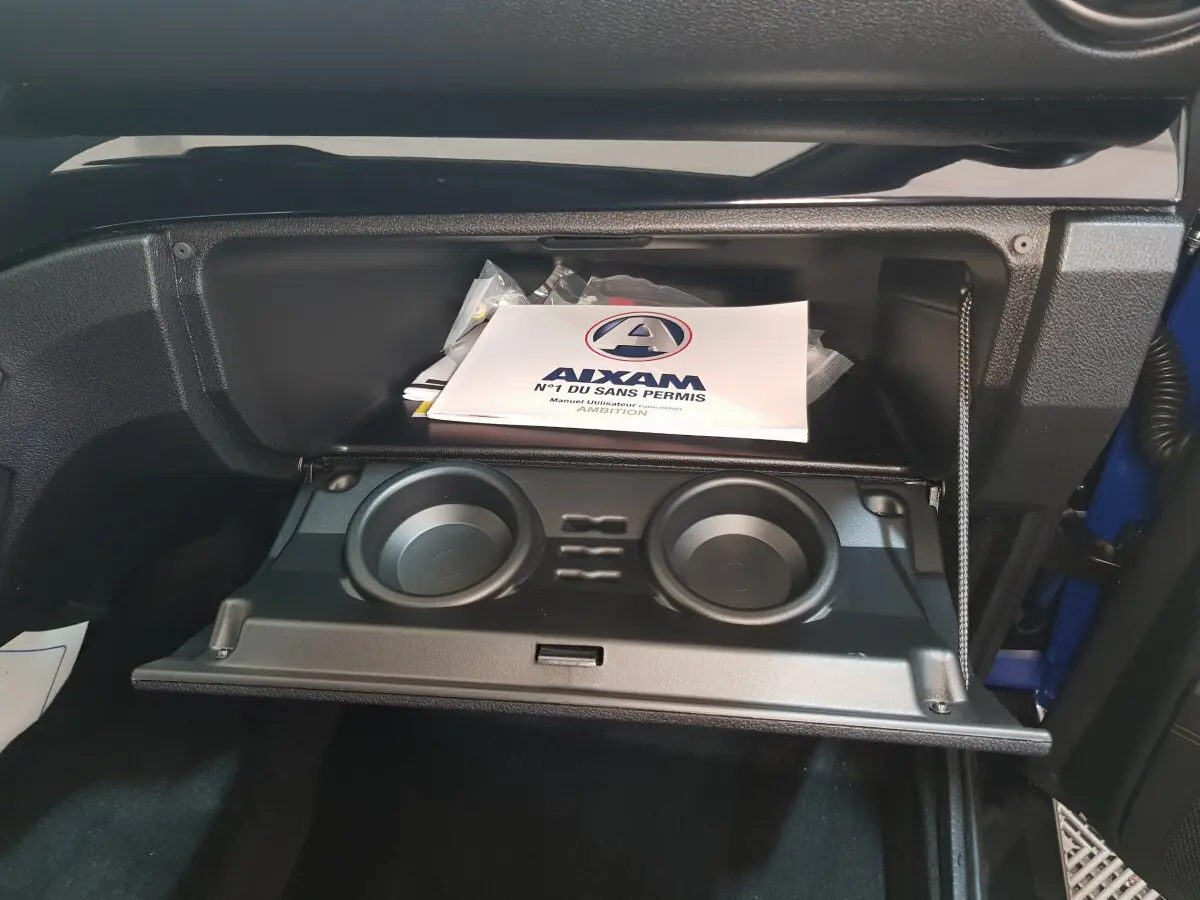 Gros plan sur la boîte à gants ouverte de l'AIXAM Ambition 0.5 City Sport bleu, avec porte-gobelets et manuel utilisateur visible.