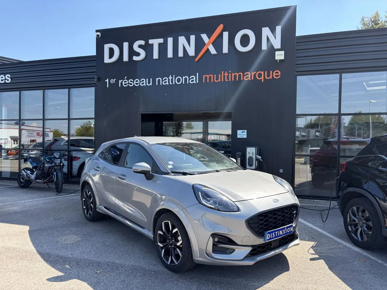 Ford Puma gris métal en 3/4 avant droit, avec jantes alliage noires et calandre ST-Line visible devant un showroom Distinxion.