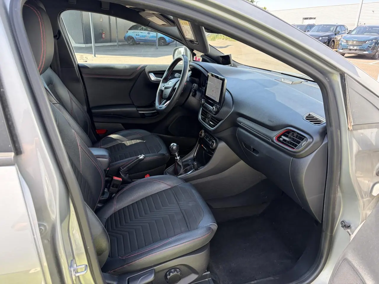 Intérieur avant du Ford Puma gris métal, vue côté conducteur, sièges noirs avec surpiqûres rouges et tableau de bord moderne.