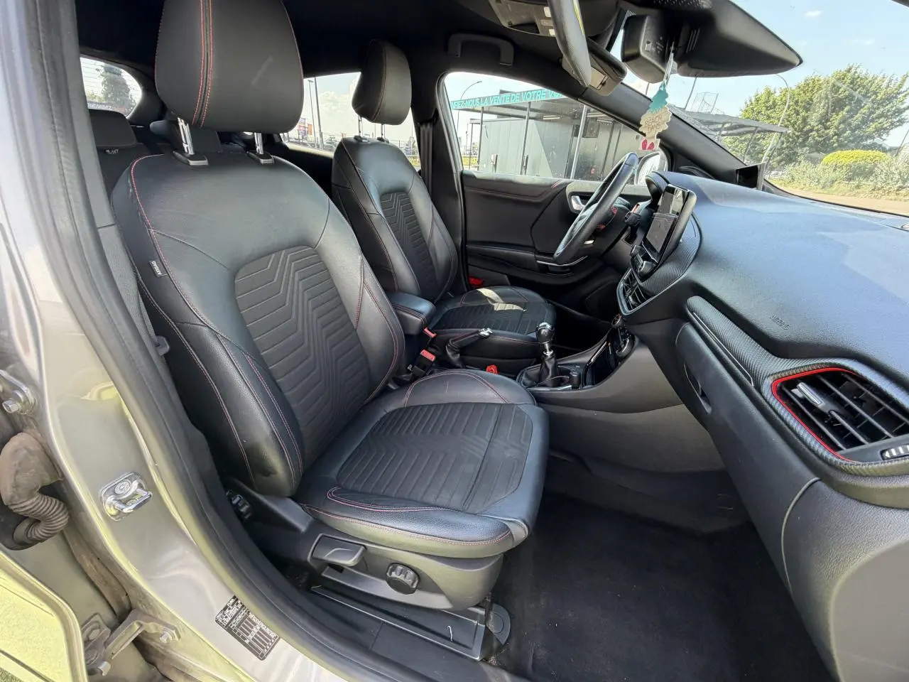 Vue intérieure côté conducteur du Ford Puma gris métal 2020, sièges noirs avec surpiqûres rouges et tableau de bord avec touches rouges.