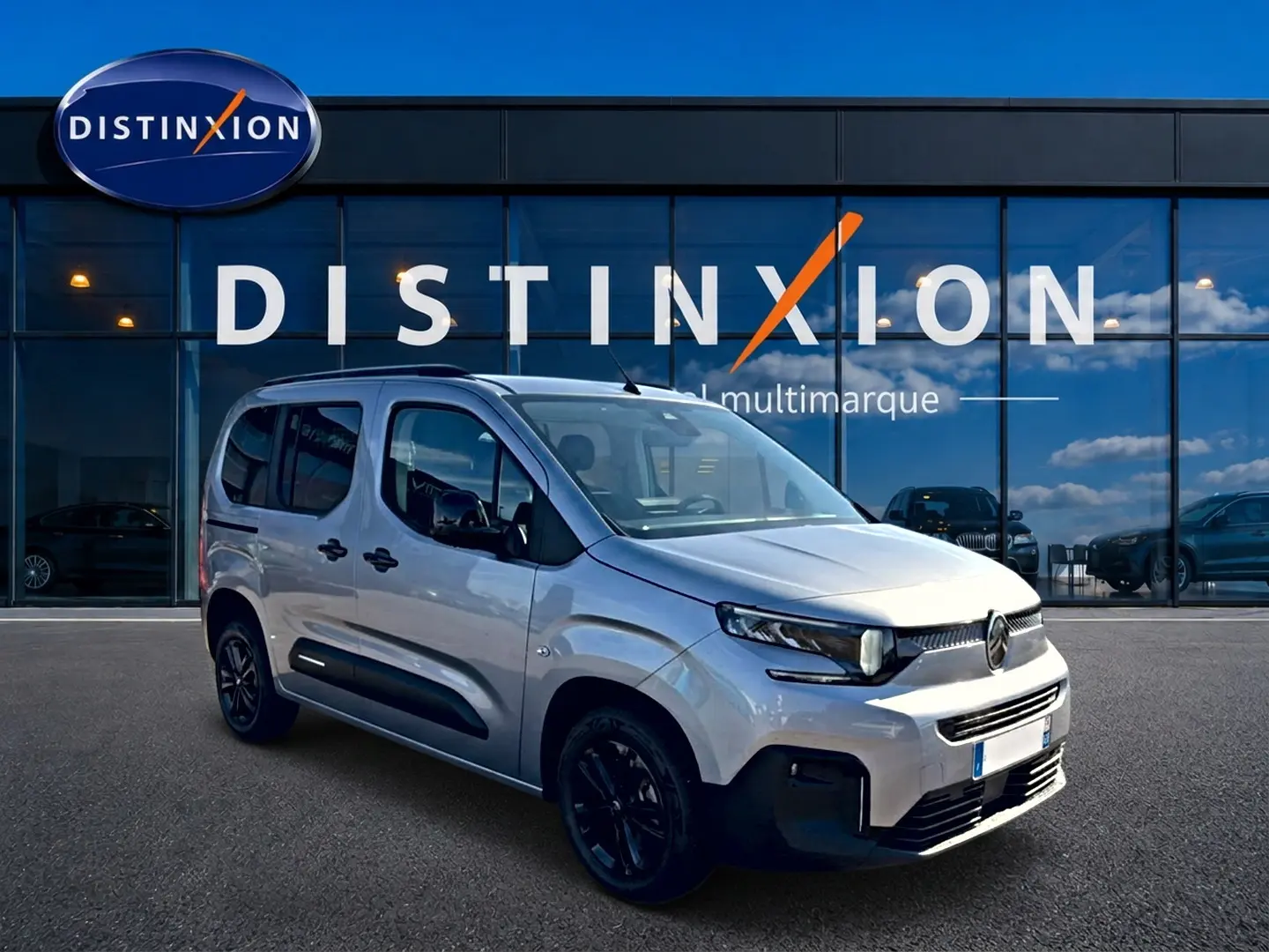 Vue 3/4 avant droit du Citroën Berlingo gris steel avec barres de toit et jantes noires devant un showroom Distinxion.
