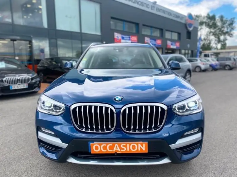 BMW X3 X-DRIVE 20 DA bleu métal vue de face avec calandre chromée et phares allumés, plaque "OCCASION" visible.