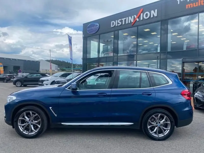 Vue latérale gauche d'un BMW X3 X-DRIVE 20 DA bleu métal, finition X-Line, garé devant un showroom Distinxion.
