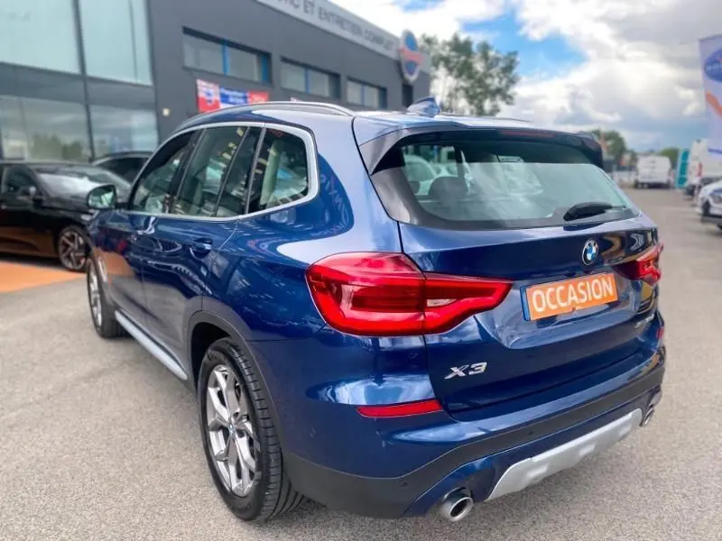 Vue 3/4 arrière droite d’un BMW X3 X-DRIVE 20 DA bleu métal avec badge X-Line et plaque occasion visible.
