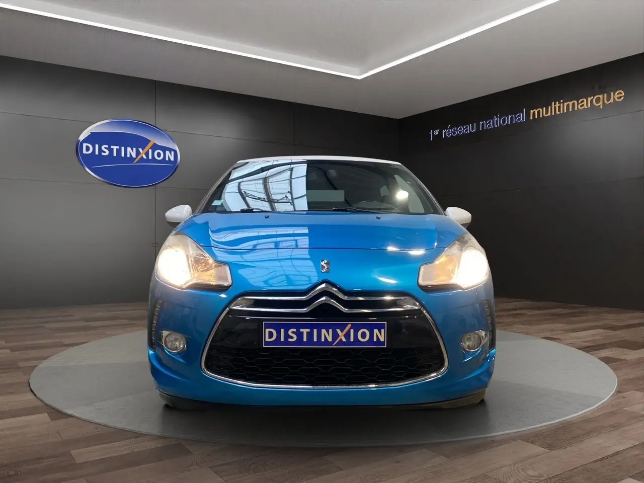Vue avant d'une Citroën DS3 bleu avec toit blanc, phares allumés, sur fond intérieur showroom Distinxion.