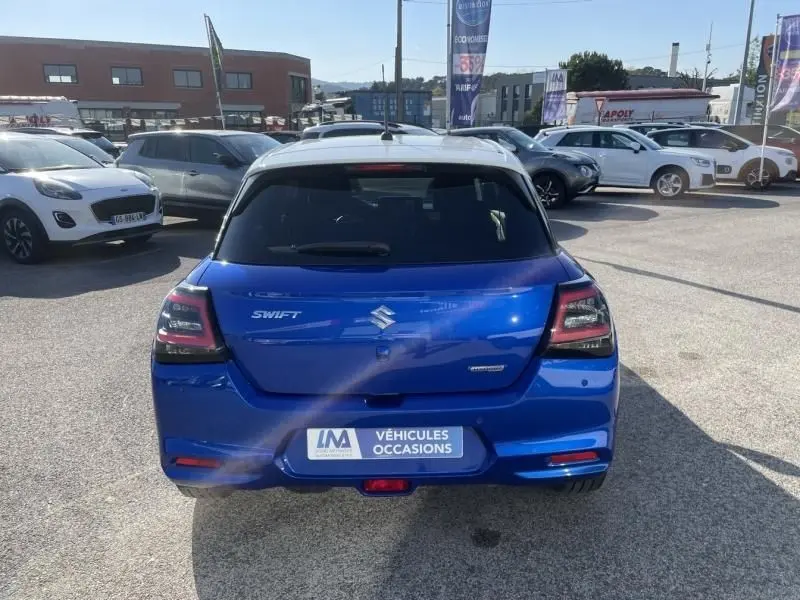 Vue arrière d'une Suzuki Swift bleu métal 2024 avec badge Hybrid et plaque "Véhicules Occasions" visible.