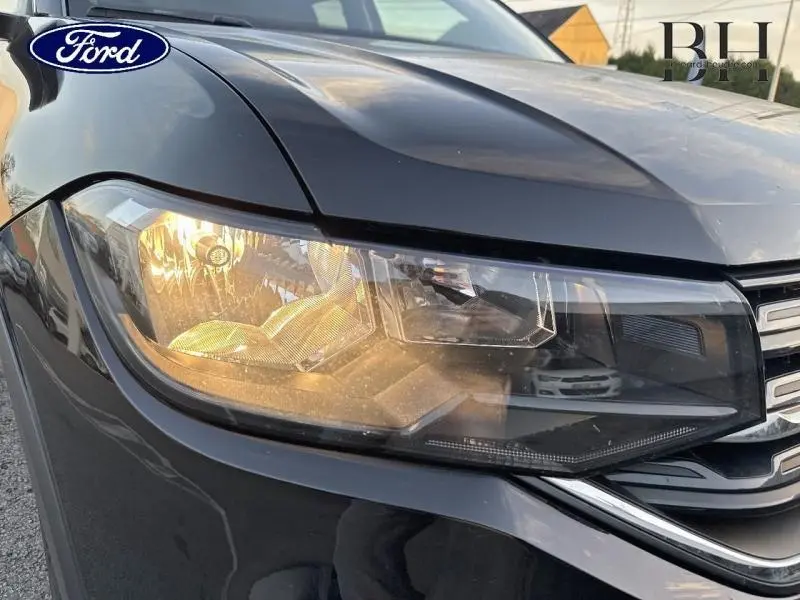 Gros plan sur le phare avant gauche d'un Volkswagen T-cross noir intense nacré, mettant en valeur ses détails lumineux.
