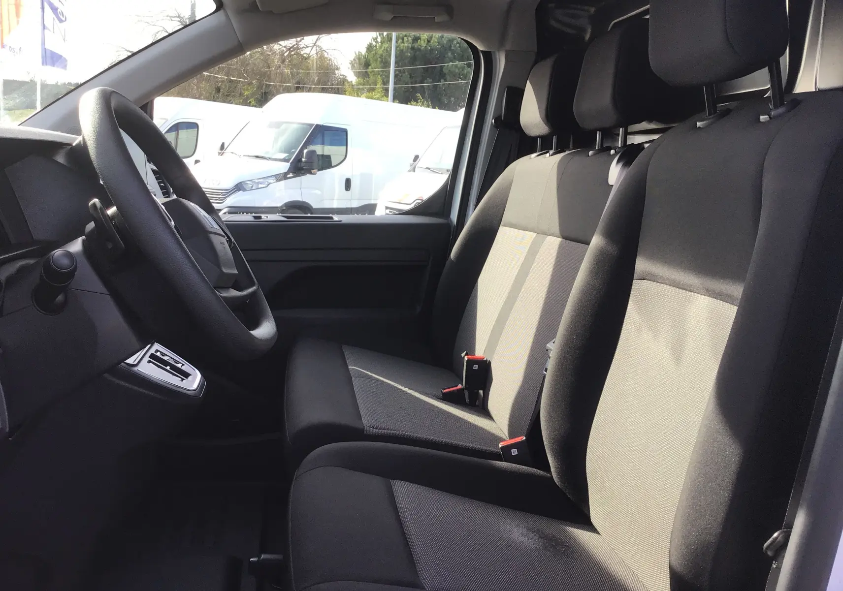 Vue intérieure côté conducteur du FIAT Scudo Fourgon blanc 2024, montrant les sièges avant en tissu noir et gris et le volant.