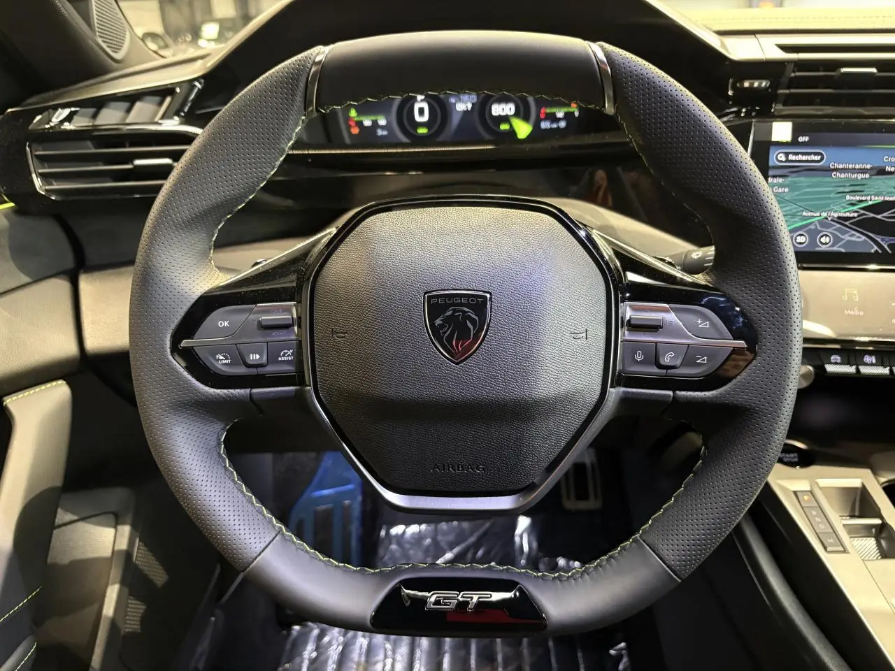 Vue rapprochée du volant GT en cuir noir avec surpiqûres vert Adamite du Peugeot 308 2026, tableau de bord digital visible.