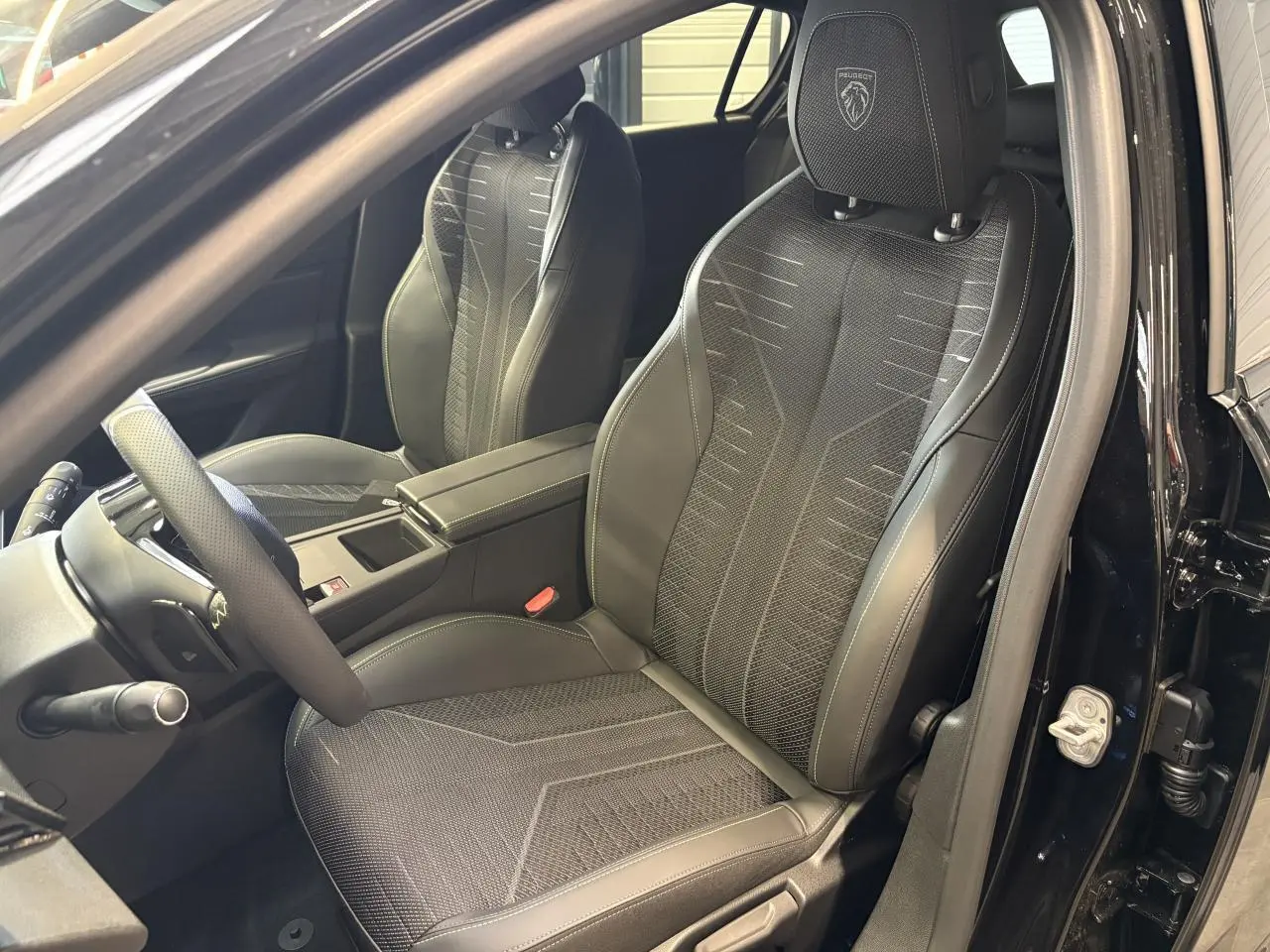 Vue intérieure côté conducteur de la Peugeot 308 GT 2026 noire métallisée, sièges sport avec logo Peugeot brodé.