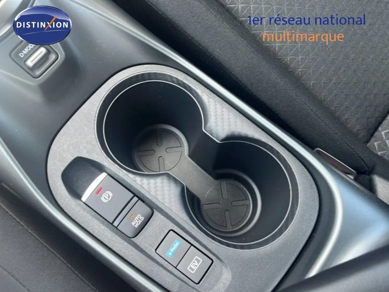 Gros plan sur la console centrale du Nissan Juke Hybrid gris, montrant les porte-gobelets et les boutons de contrôle électrique.