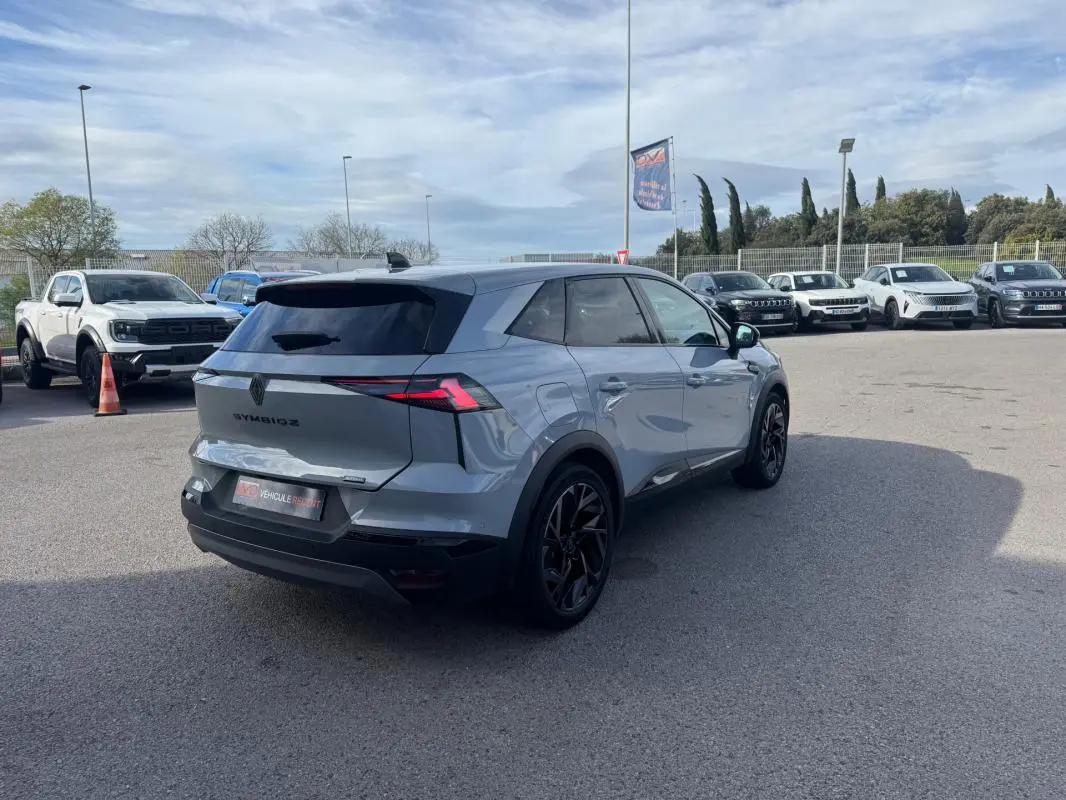 Vue 3/4 arrière droite d'un Renault SYMBIOZ gris clair avec jantes alliage noires et antenne requin sur parking.