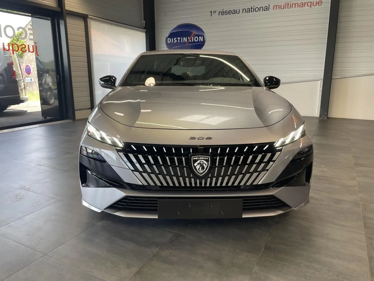Vue frontale d'une Peugeot 308 gris Artense avec calandre noire brillante et logo lion illuminé en showroom.