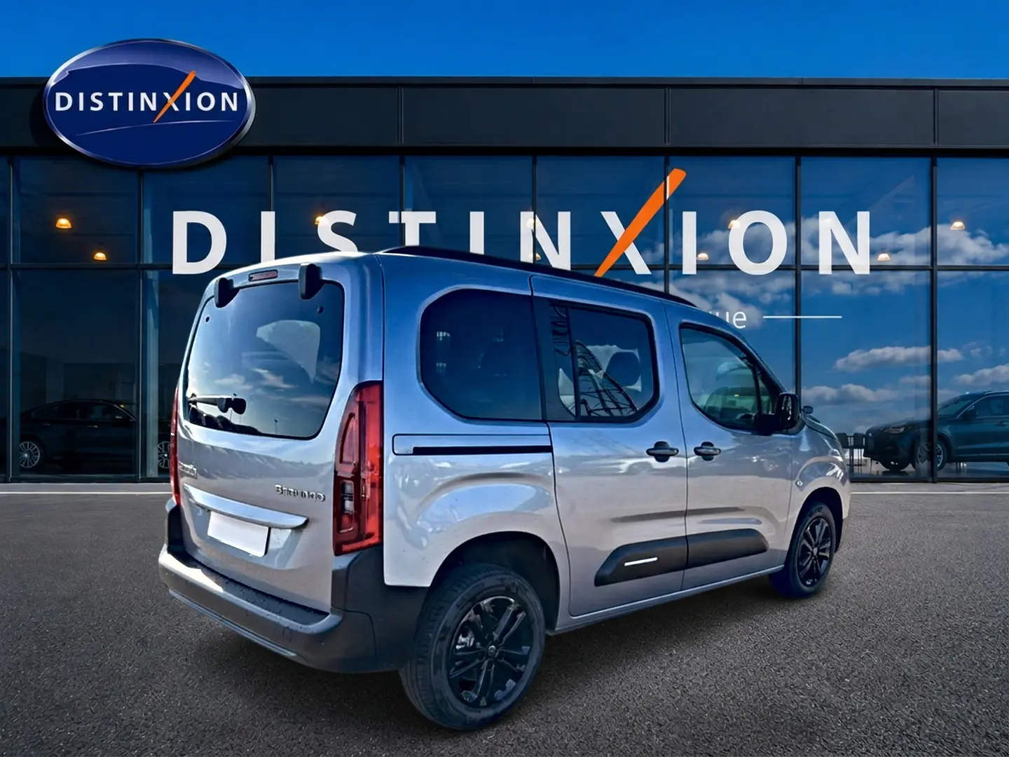 Vue 3/4 arrière droite du Citroën Berlingo Steel Grey avec barres de toit et jantes alu noires devant un showroom Distinxion.