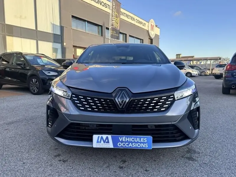 Vue frontale d'une Renault Clio V gris shiste 2025 avec calandre noire et logo Renault central, sur parking extérieur.