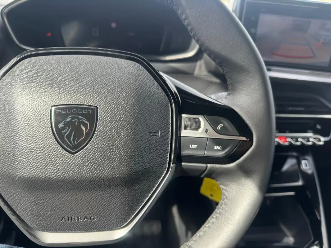 Vue rapprochée du volant noir de la Peugeot 2008 2024 avec commandes intégrées et tableau de bord numérique en arrière-plan.