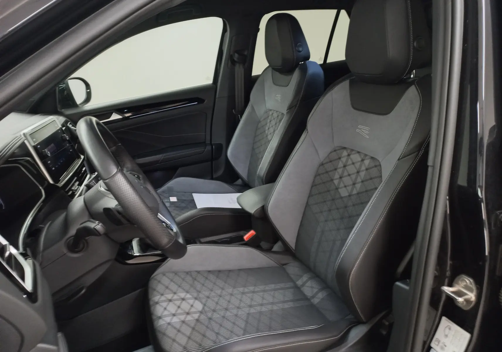 Vue intérieure côté conducteur du Volkswagen T-Roc 1.5 TSI R-Line 2025, sièges sport en cuir et alcantara noirs avec surpiqûres grises.