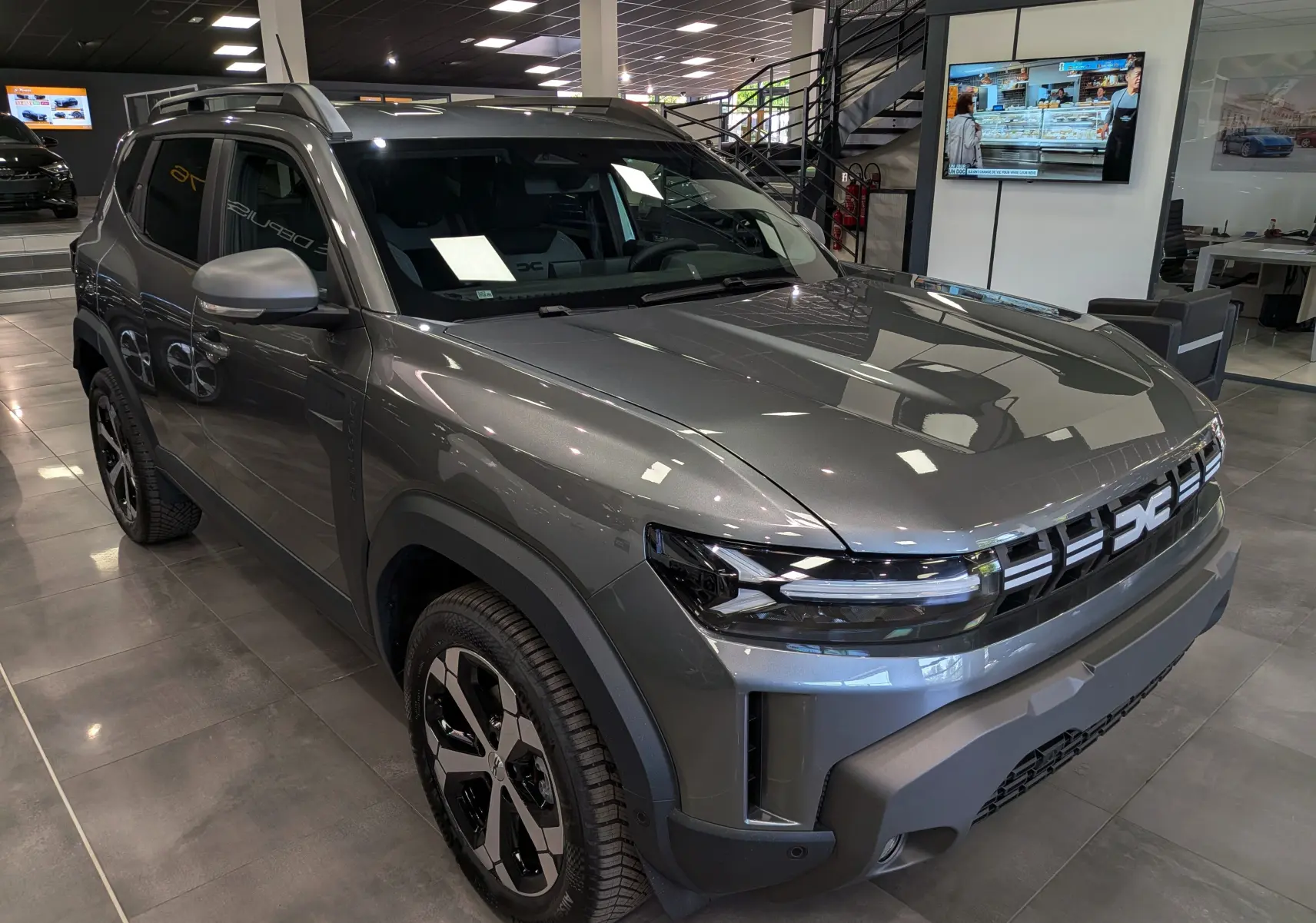 Vue 3/4 avant du Dacia Duster Hybrid-G 150 CH 4x4 gris schiste, avec calandre moderne et jantes bi-ton en showroom.