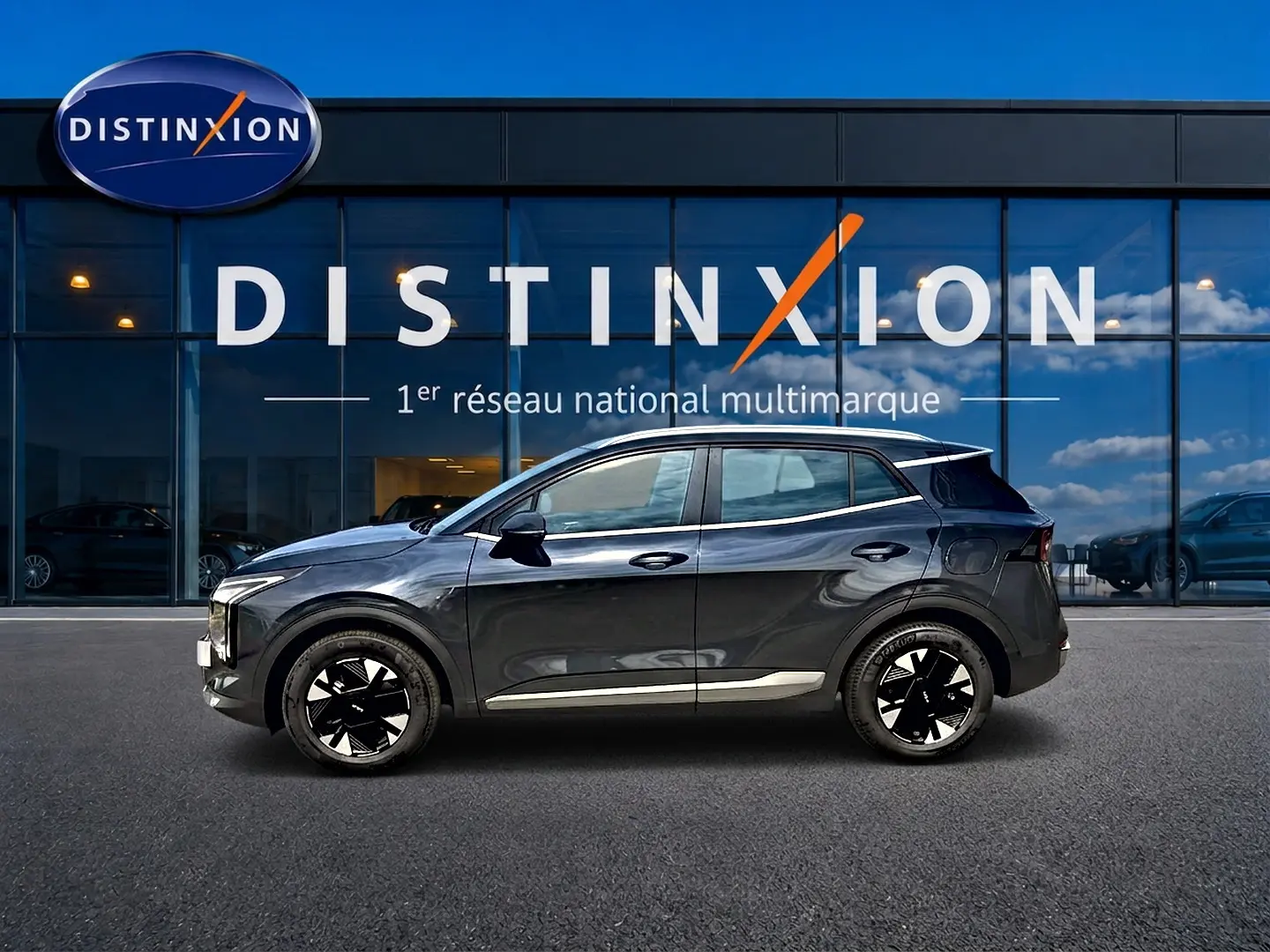 Profil côté gauche du KIA SPORTAGE 1.6 hybride 239 gris eclipse devant une vitrine avec logo Distinxion.