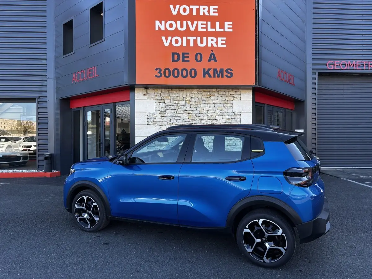 Vue latérale droite d'une Citroën C3 100 BVM PLUS bleu avec toit noir et jantes distinctives, devant un bâtiment commercial.