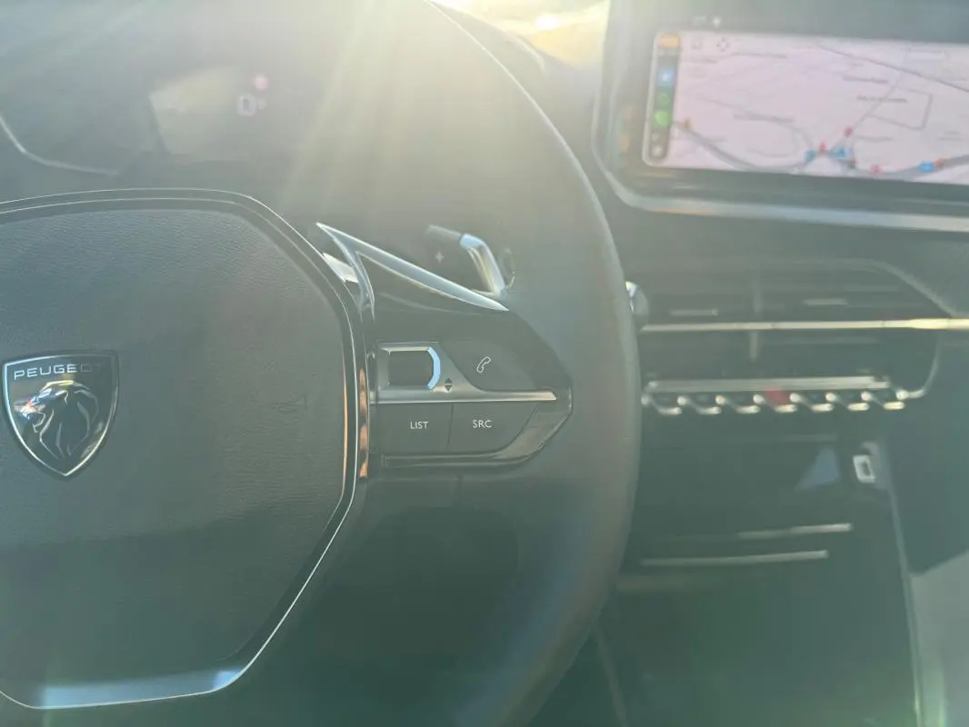 Vue rapprochée du volant noir du Peugeot 2008 Hybrid 136 gris avec commandes et écran tactile en arrière-plan.