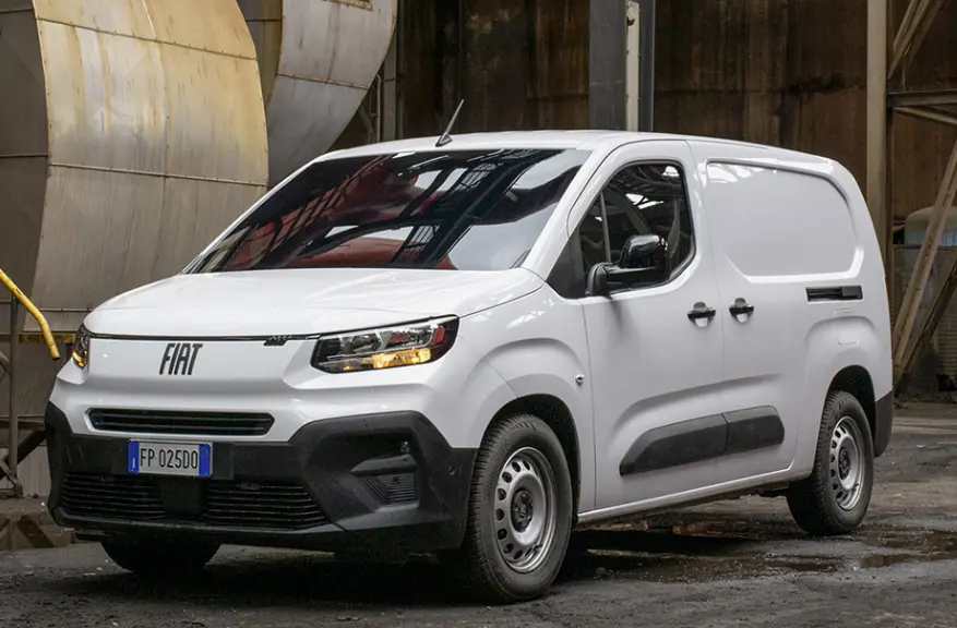 Fiat Doblo fourgon blanc vu en 3/4 avant droit dans un environnement industriel, avec calandre et phares allumés.