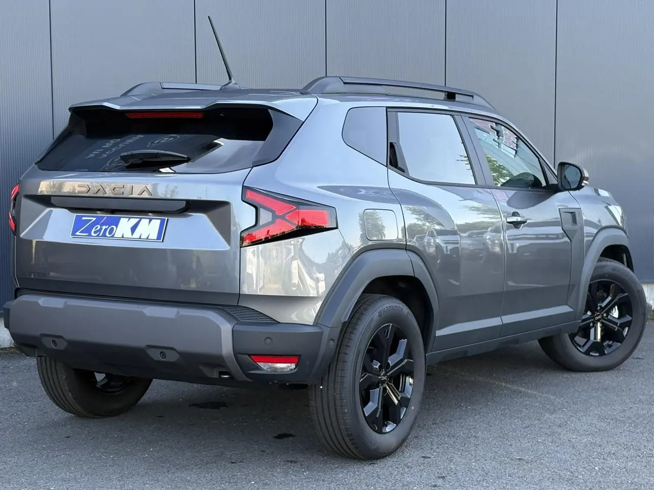 Vue 3/4 arrière droit du Dacia Duster 2026 gris schiste avec jantes alliage noires et barres de toit longitudinales.
