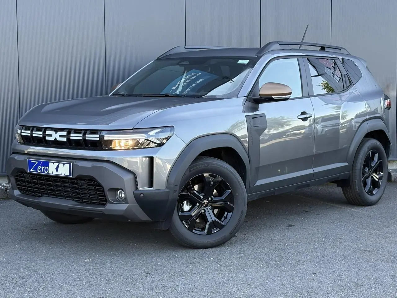Vue 3/4 avant droit du Dacia Duster 2026 gris schiste avec jantes noires et barres de toit noires.