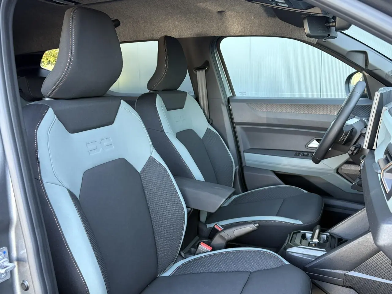 Vue intérieure côté conducteur du Dacia Duster 2026 gris Schiste, sièges bi-ton gris clair et noir avec accoudoir central.