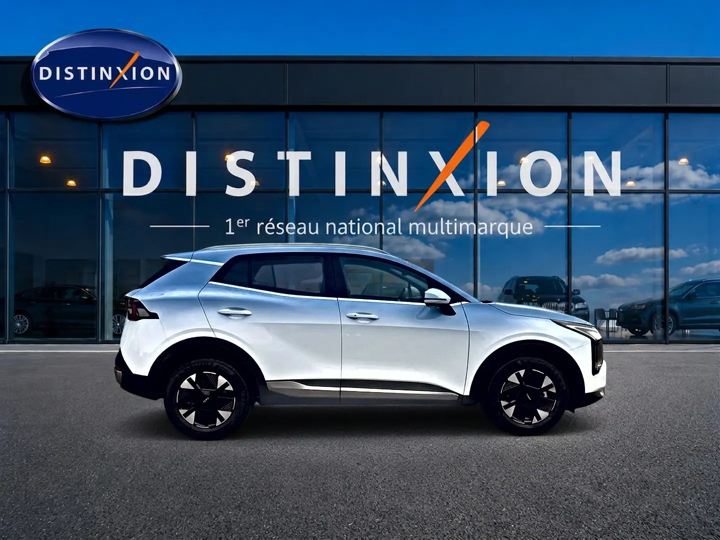 Profil droit d’un KIA SPORTAGE 1.6 hybride blanc 2026 avec jantes alu noires devant une vitrine Distinxion.