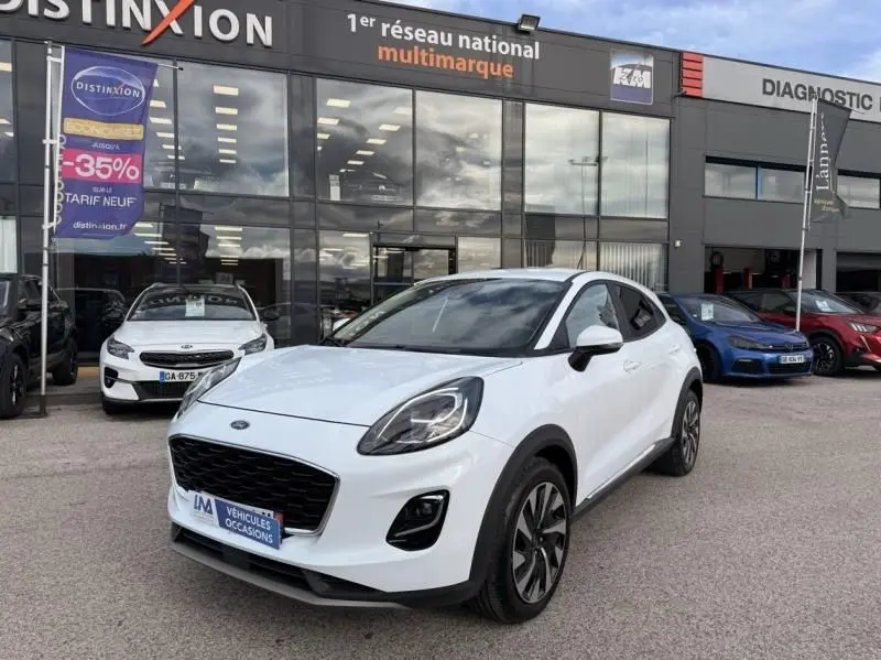 Ford Puma blanc en 3/4 avant droit, avec calandre noire et jantes alliage, devant un concessionnaire multimarque.