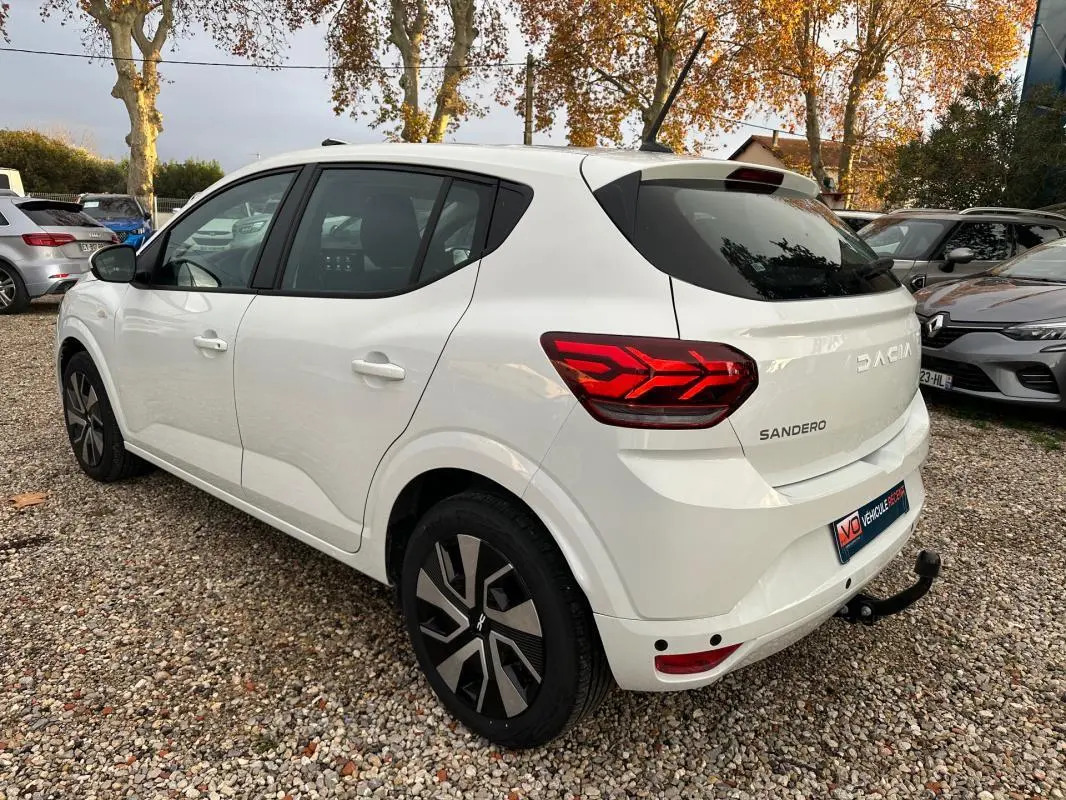Vue 3/4 arrière droite d'une Dacia Sandero blanche 2024 avec feux arrière LED et attelage de remorque visible.