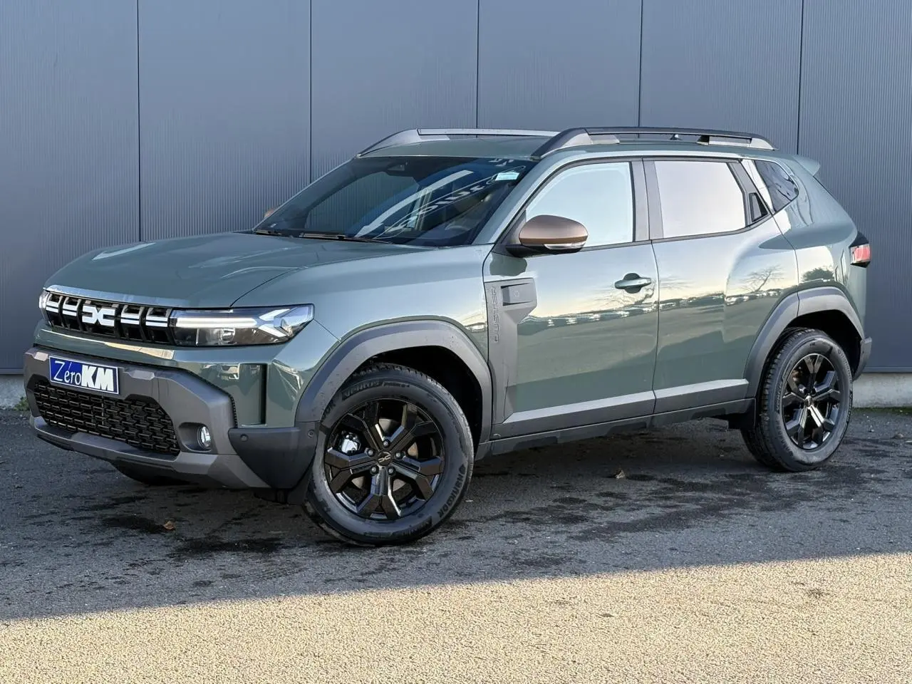 Vue 3/4 avant droit du Dacia Duster 2026 kaki lichen avec jantes noires et barres de toit noires.