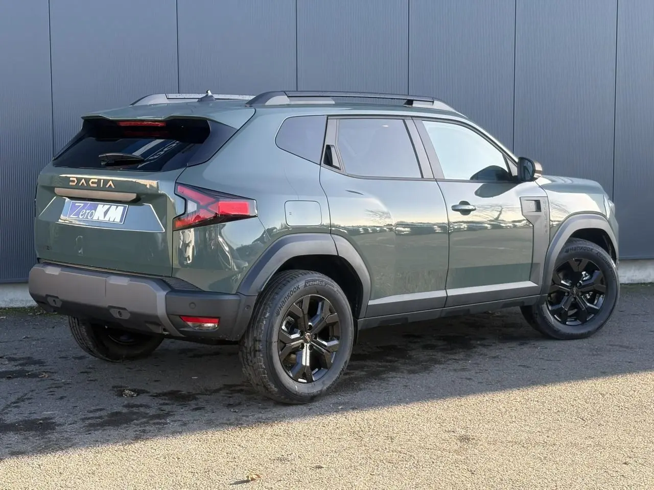 Vue 3/4 arrière droite du Dacia Duster 2026 kaki lichen avec jantes alliage noires et barres de toit longitudinales.