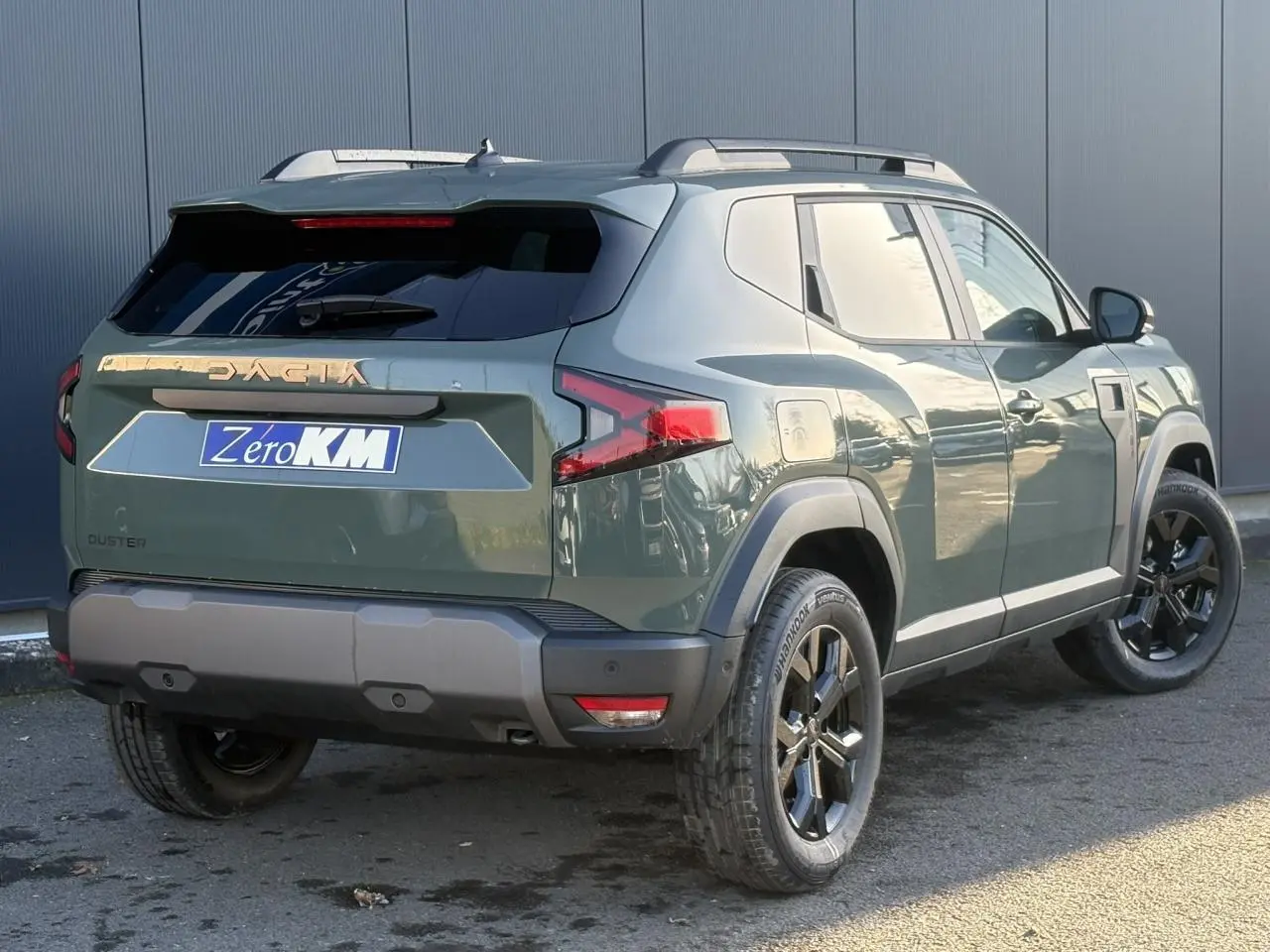 Vue 3/4 arrière droite du Dacia Duster 2026 kaki lichen avec jantes noires et barres de toit longitudinales.