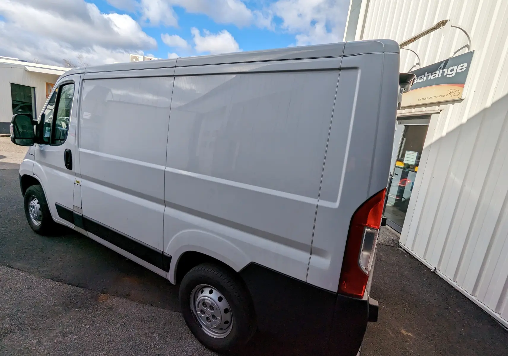 Fourgon blanc FIAT Ducato vu en 3/4 arrière côté droit, stationné devant un bâtiment industriel.