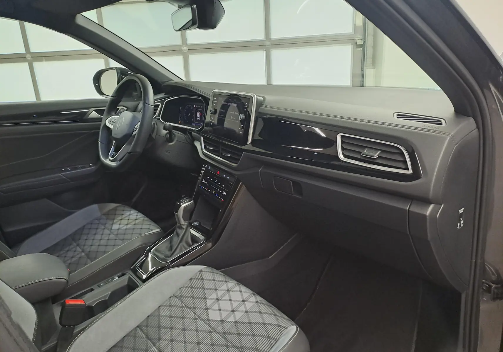 Vue intérieure côté passager du Volkswagen T-Roc 2025, tableau de bord noir avec sièges tissu gris à motif géométrique.