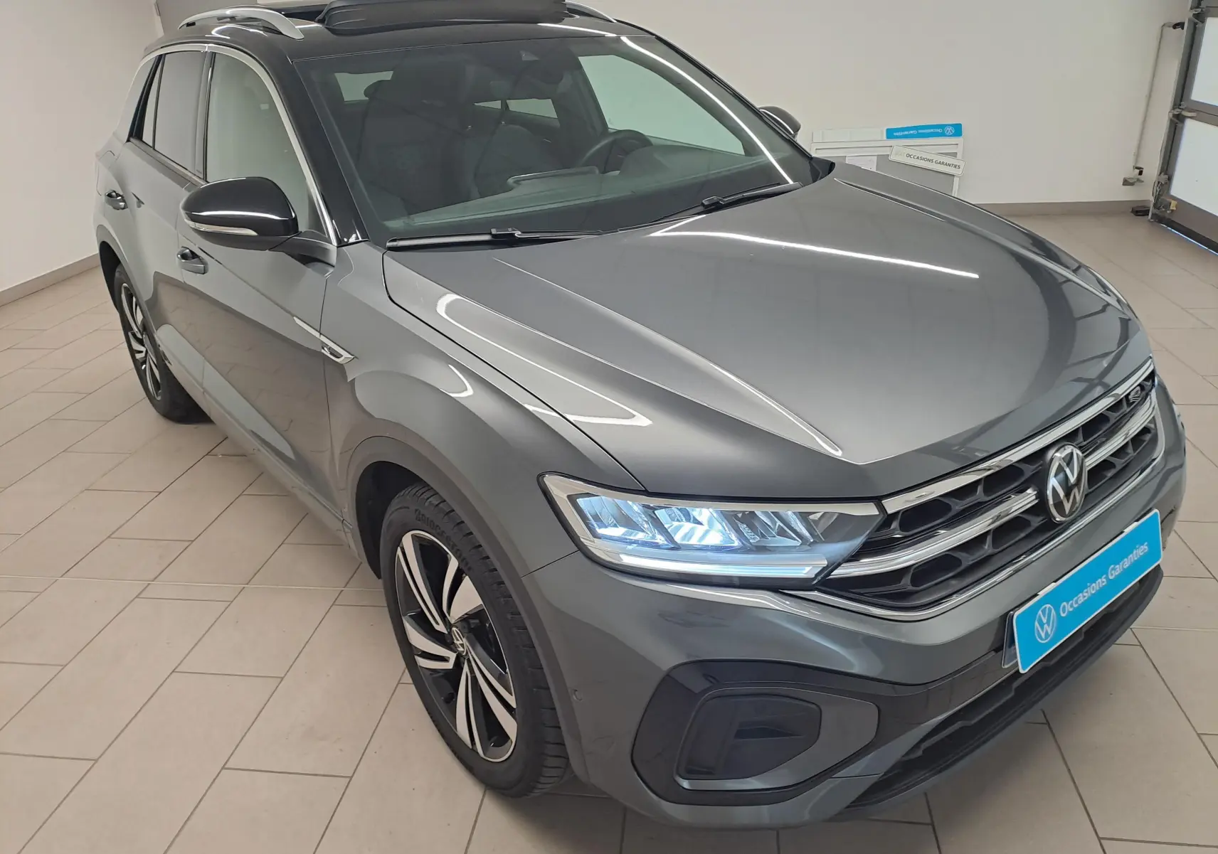 Vue 3/4 avant droite d’un Volkswagen T-Roc 2025 gris Indium avec toit noir et phares LED allumés dans un showroom.