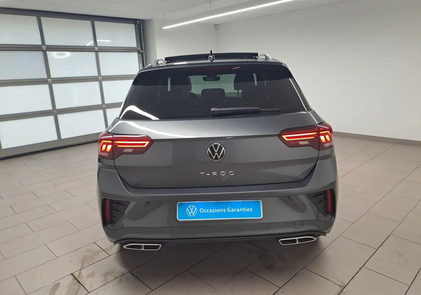 Vue arrière d'un Volkswagen T-Roc gris Indium avec toit noir et feux LED allumés dans un garage.