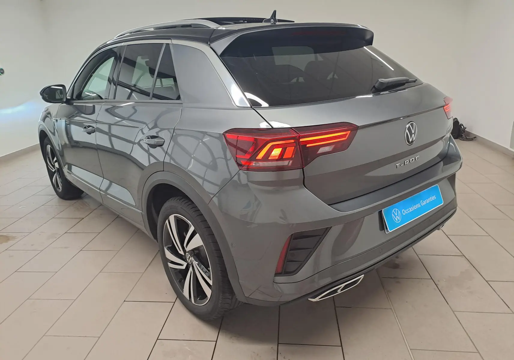 Vue 3/4 arrière droite du Volkswagen T-Roc 2025 gris Indium avec toit noir et feux arrière LED allumés.
