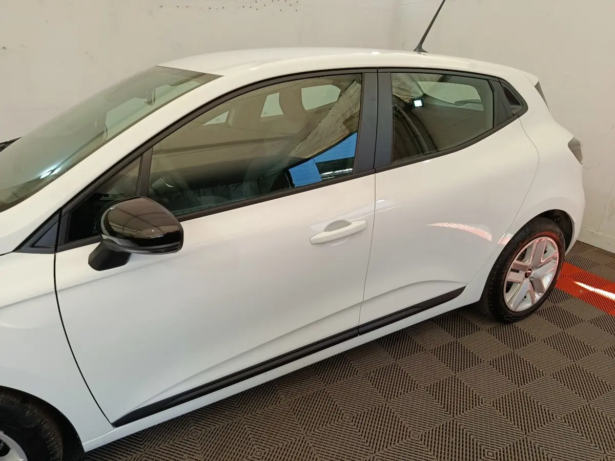 Profil côté gauche de la Renault Clio blanche 2024 avec rétroviseur noir et jantes alliage visibles en intérieur neutre.
