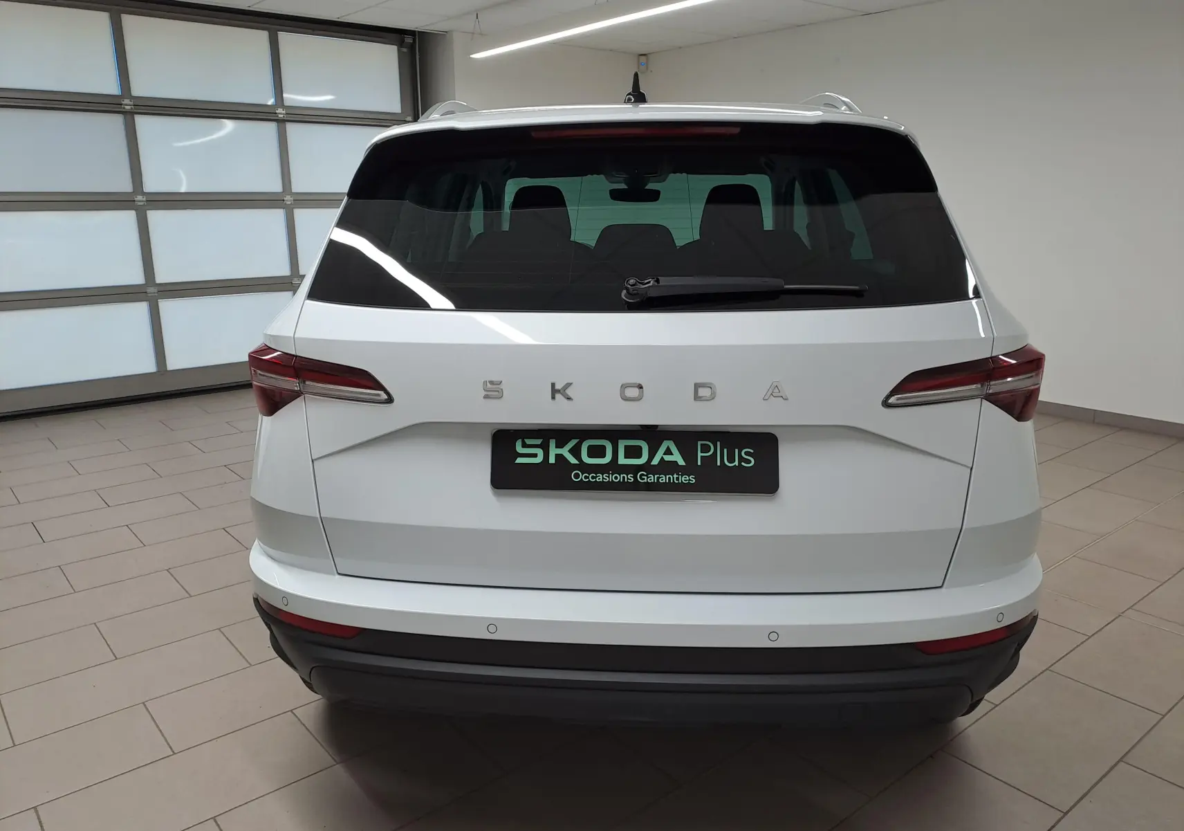 Vue arrière d'un Skoda Karoq blanc lune 2025 avec feux arrière LED et entourage de vitres chromé en intérieur showroom.