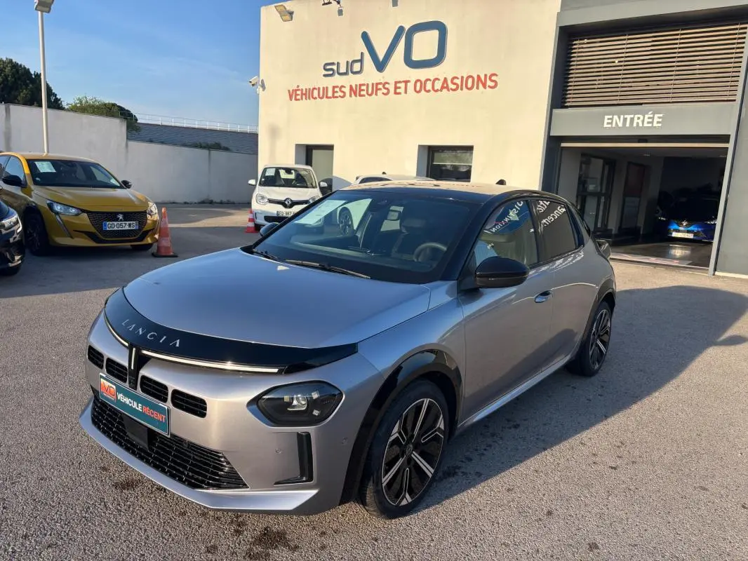 Lancia Ypsilon gris vue 3/4 avant droit, avec jantes 17 pouces et calandre noire brillante, stationnée devant un garage.