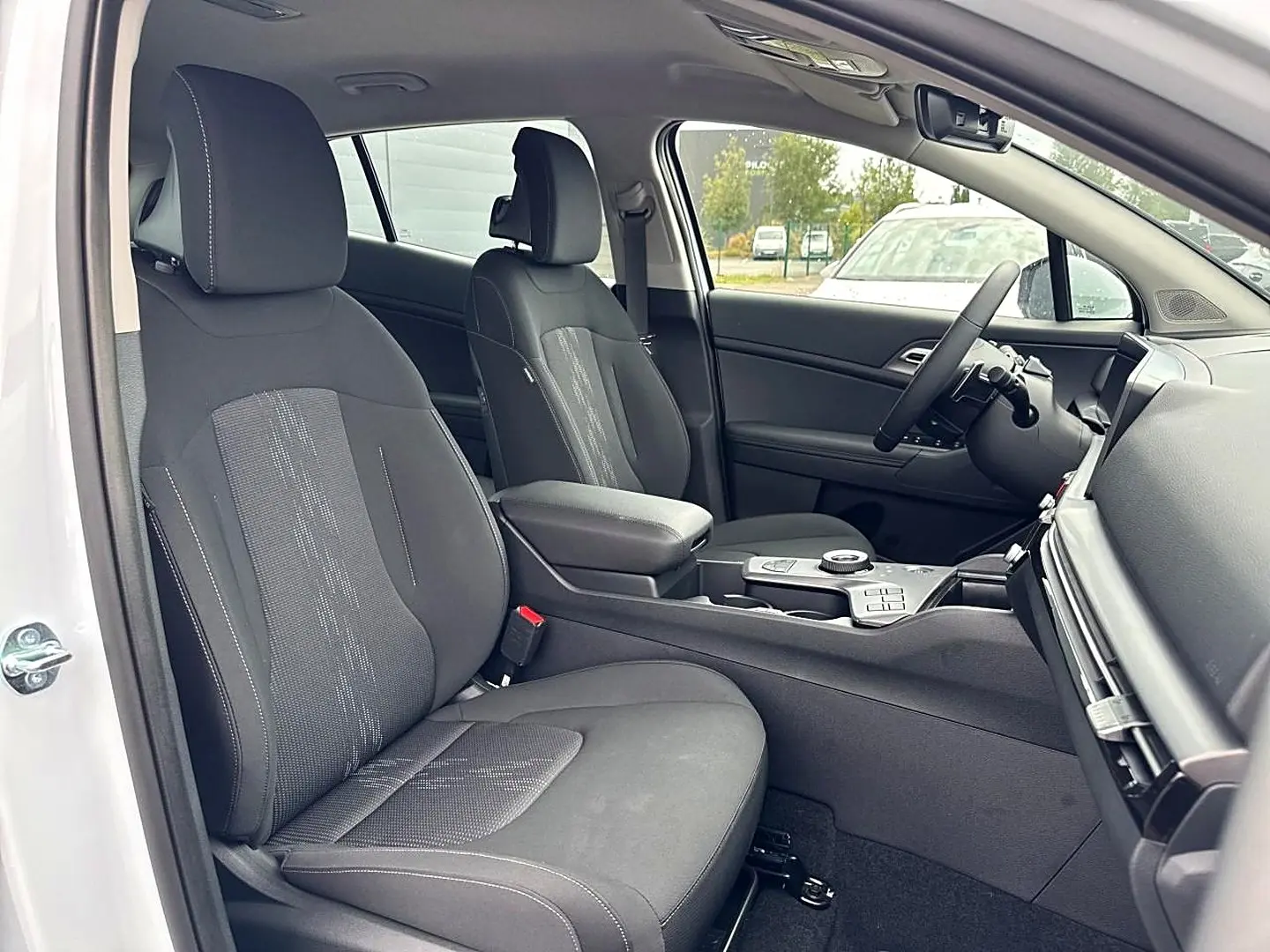 Intérieur avant du Kia Sportage blanc 2026, vue côté conducteur montrant sièges noirs et console centrale avec accoudoir.