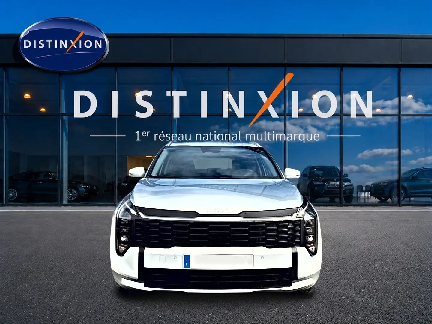 Vue frontale d'un Kia Sportage 1.6 hybride blanc avec calandre noire et feux LED verticaux, devant un showroom Distinxion.