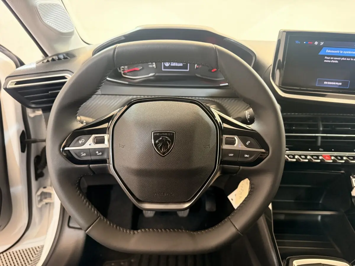 Vue rapprochée du volant cuir noir de la Peugeot 208 blanche, avec tableau de bord et écran tactile central visibles.