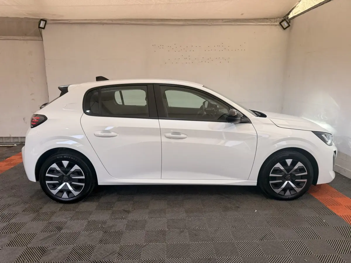 Profil droit d'une Peugeot 208 blanche 2025 avec jantes alliage et vitres teintées dans un garage.