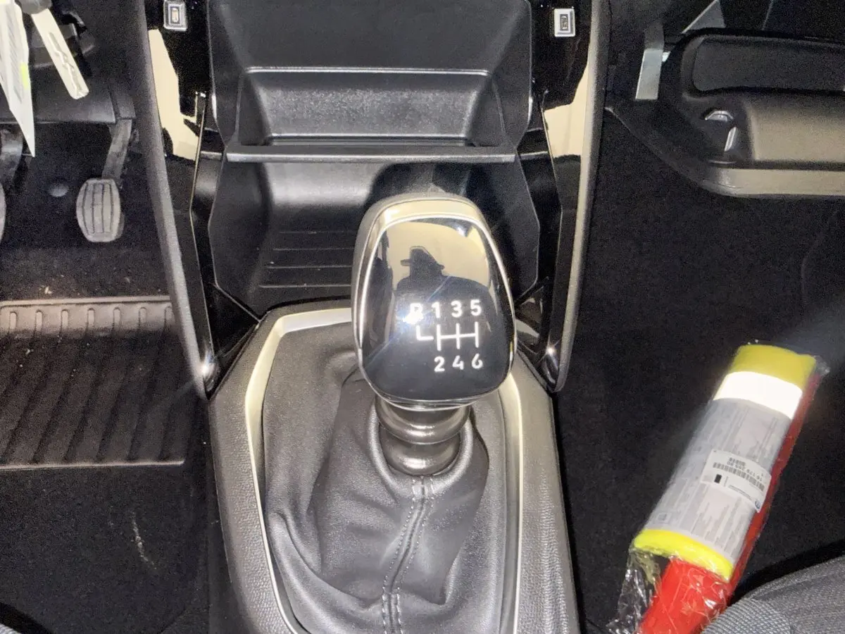Gros plan sur le levier de vitesse manuel noir dans l’habitacle d’une Peugeot 208 blanche, avec pédales et extincteur visibles.
